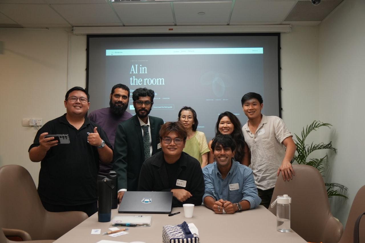 Participants and hosts of the AI in the room workshop grouped in front of the projected title, Singapore, 14 September 2026