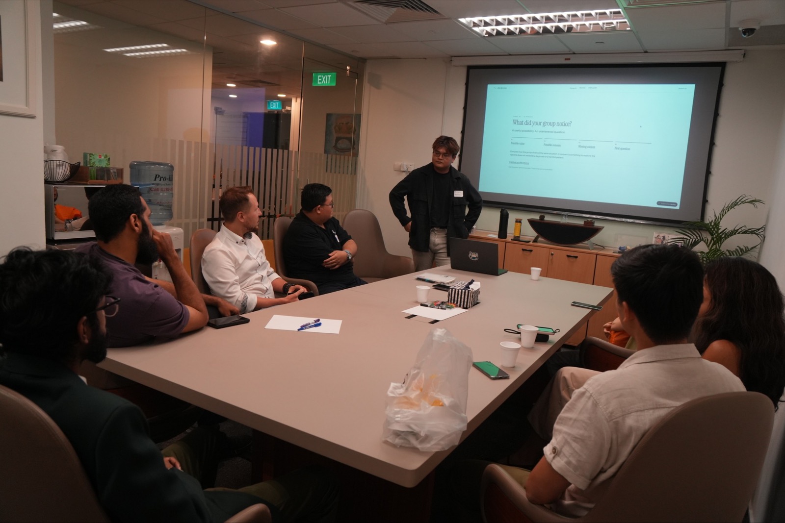 Facilitator standing by a projection screen while workshop participants discuss in small groups around a table